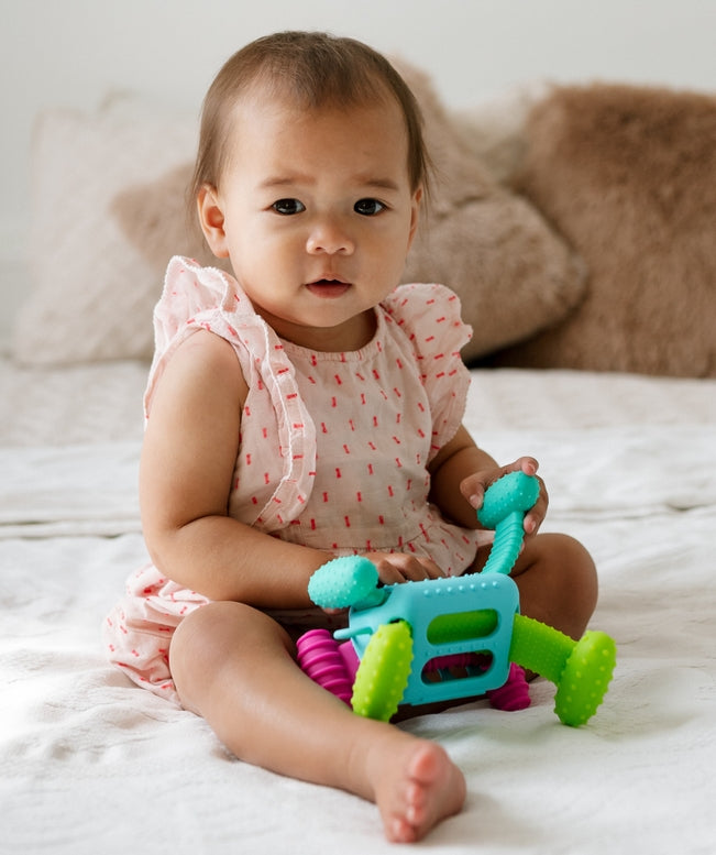An infant sitting upright on a bed, playing with the Knudll Sensory Activity Toy by Mobi Games.