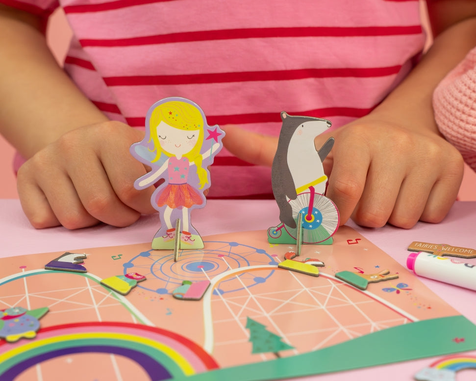 Child playing with the Floss & Rock Magnetic Scene & Doodle Set in the Rainbow Fairy theme, setting up magnetic characters like a fairy and a seal on a bicycle on a colorful magnetic board.