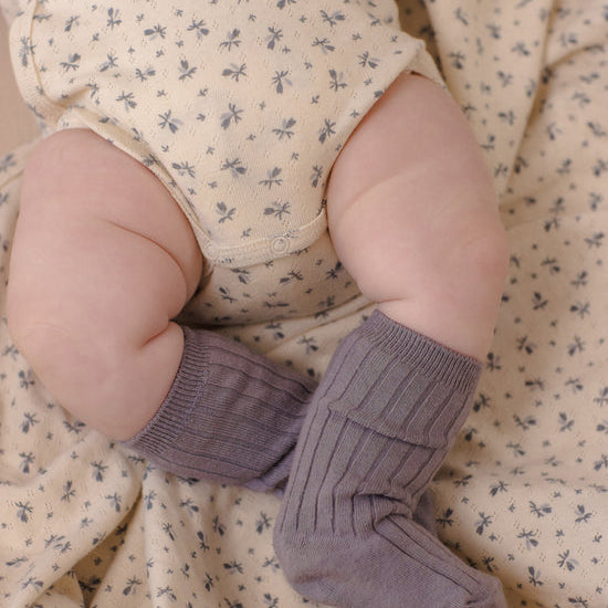 A close-up of a baby's legs wearing ribbed mauve-purple knit booties, resting on a cream-colored floral blanket.