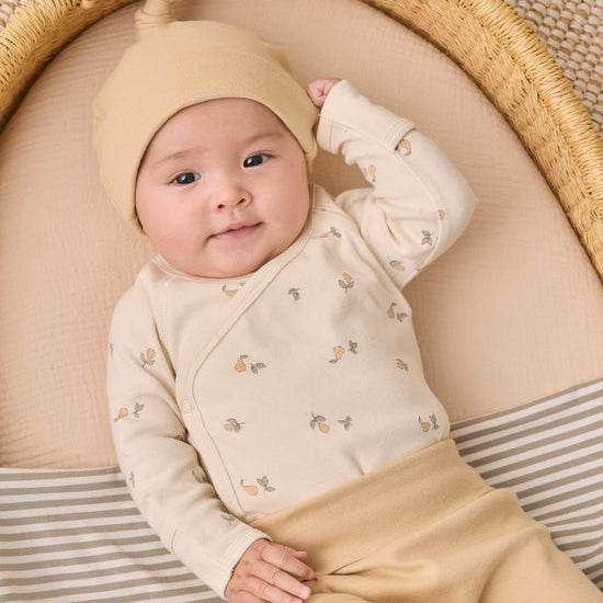 A baby lying on their back inside a woven moses basket, wearing a cream-colored long-sleeve wrap bodysuit with a small floral print, a matching cream knit hat, and tan pants.