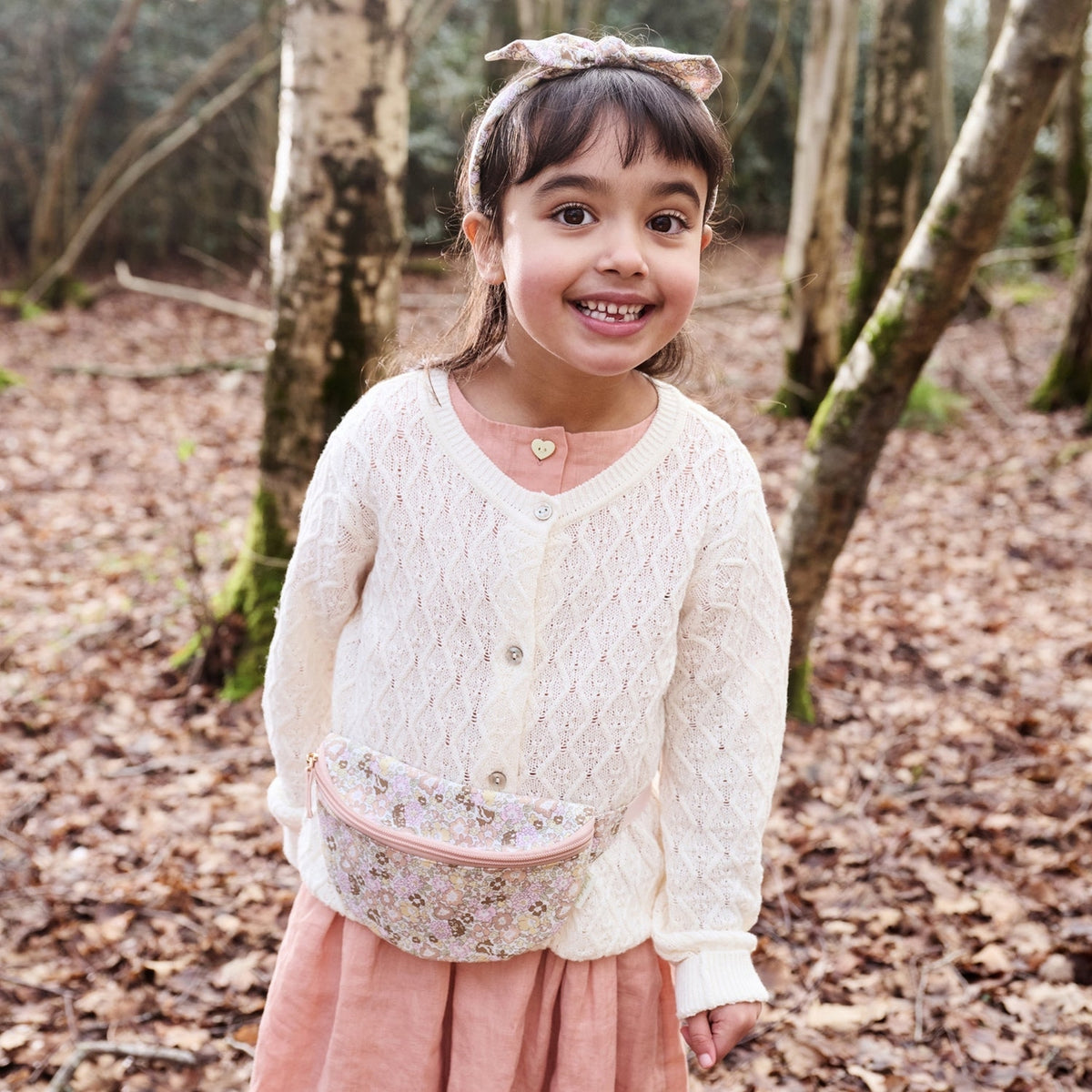 Young girl in a forest wearing a white lace cardigan and pink skirt with a floral bag.