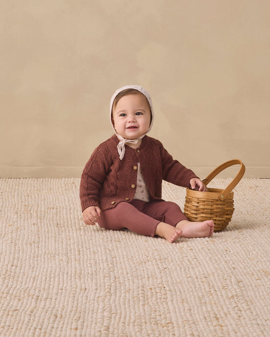A baby sitting on a textured cream rug against a warm beige wall, wearing a brown knit cardigan, matching brown pants, a white bonnet, and holding a small woven basket.