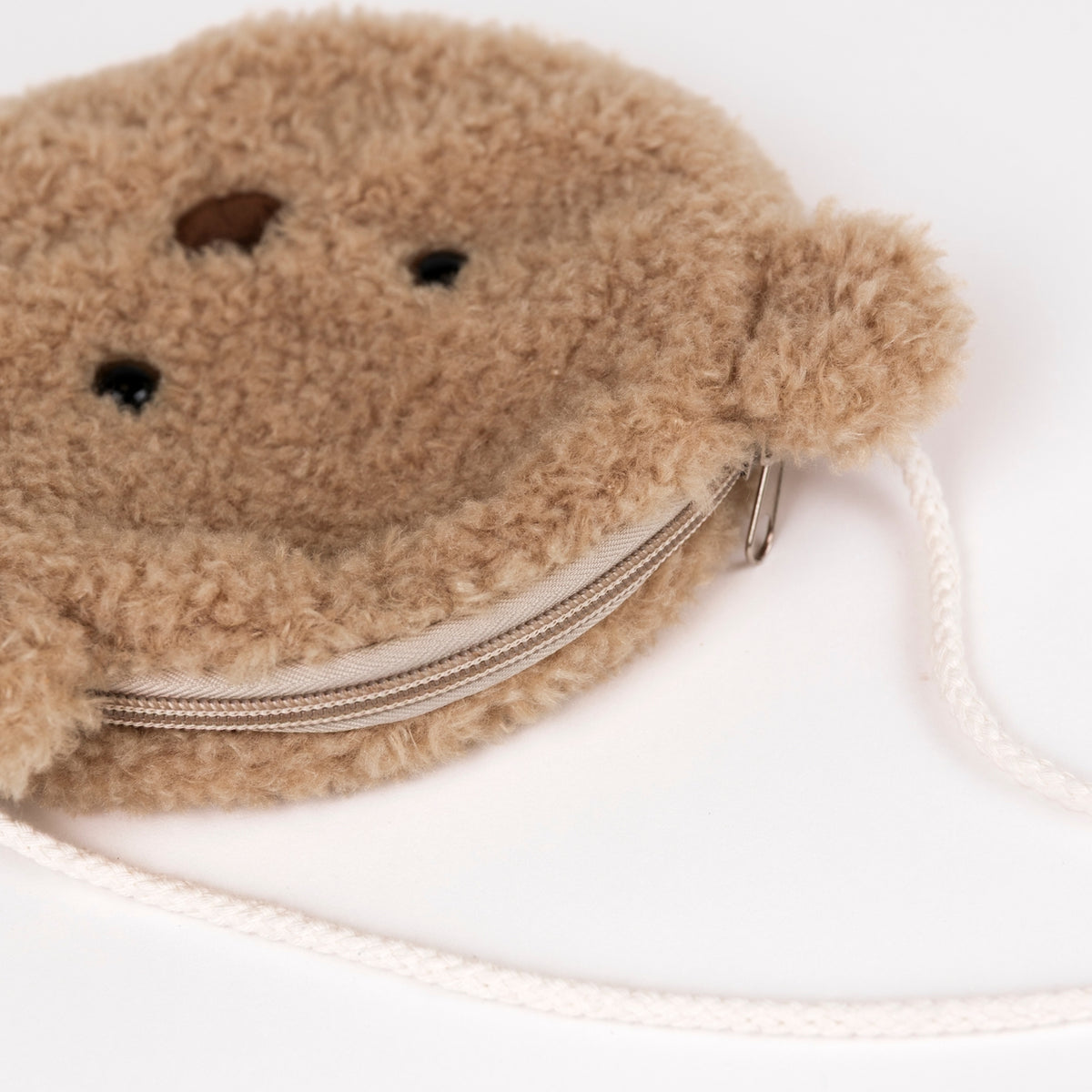A close-up of a round, plush teddy bear face zipper pouch in light brown/tan faux fur, featuring embroidered eyes and nose, a silver zipper, and a cream-colored braided strap on a white background.