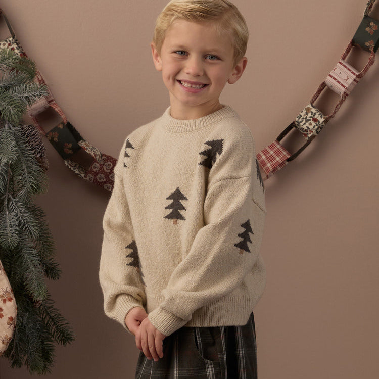 A young blonde boy smiling while wearing a cream knit sweater with black pine tree motifs and dark plaid pants, standing against a warm brown wall with a holiday garland in the background.