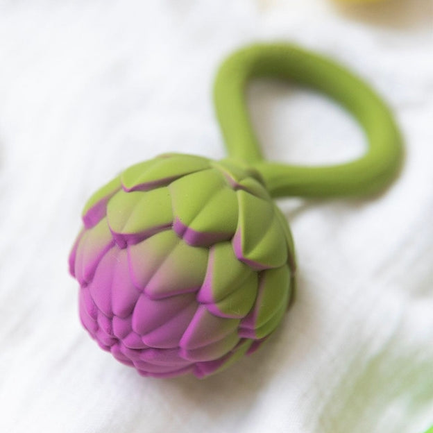 Oli & Carol Artichoke Rattle Toy resting on a light surface, highlighting the textured purple-tipped bulb and integrated green ring handle.