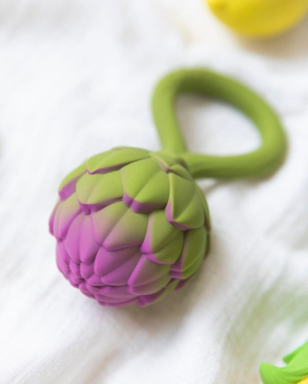 Oli & Carol Artichoke Rattle Toy resting on a light surface, highlighting the textured purple-tipped bulb and integrated green ring handle.
