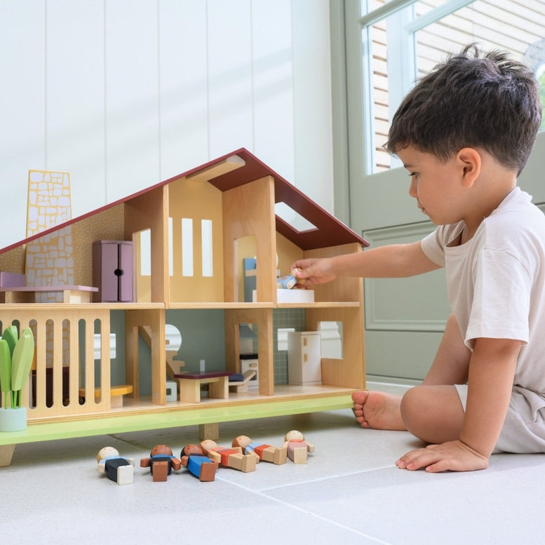 A young child sits on the floor playing with the Wooden Cedar Chalet Modernist Dollhouse by Mentari, placing miniature figures and furniture inside the open-front mid-century modern home while additional wooden dolls and pieces rest on the floor nearby.