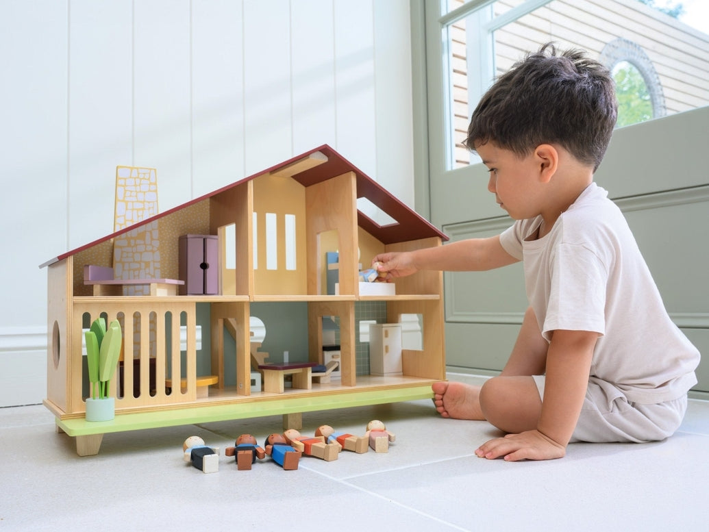 A young child sits on the floor playing with the Wooden Cedar Chalet Modernist Dollhouse by Mentari, placing miniature figures and furniture inside the open-front mid-century modern home while additional wooden dolls and pieces rest on the floor nearby.
