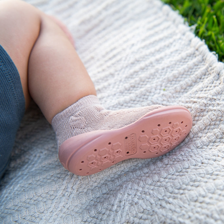 A close-up of a child's foot wearing a light pink knit sock and a matching pink shoe with a honeycomb-patterned sole, resting on a textured cream blanket with green grass visible in the background.