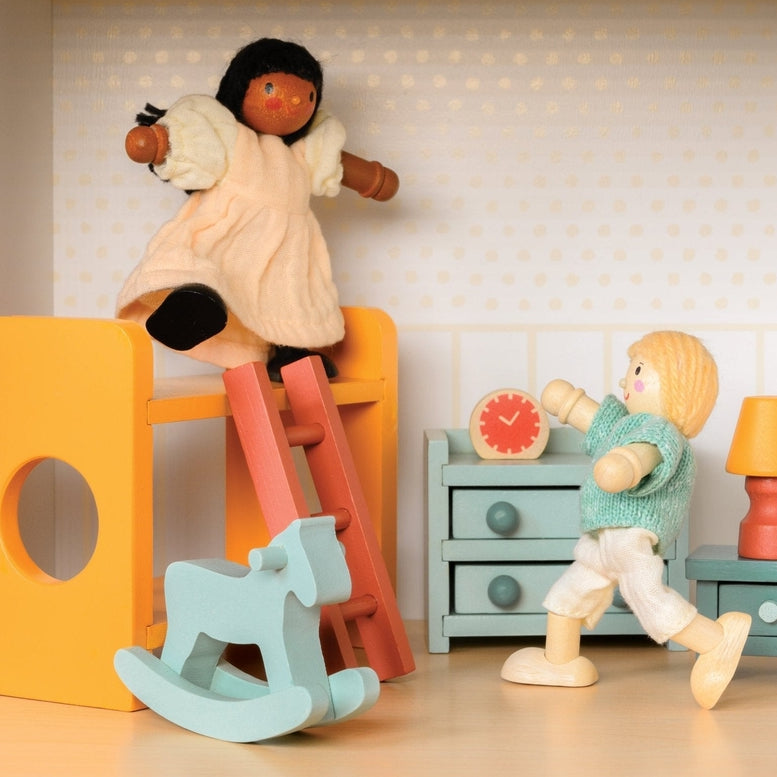Wooden doll figure climbing down the ladder of a yellow bunk bed in a miniature room setting with a slide, light blue rocking horse, chest of drawers, and bedside table with a lamp.