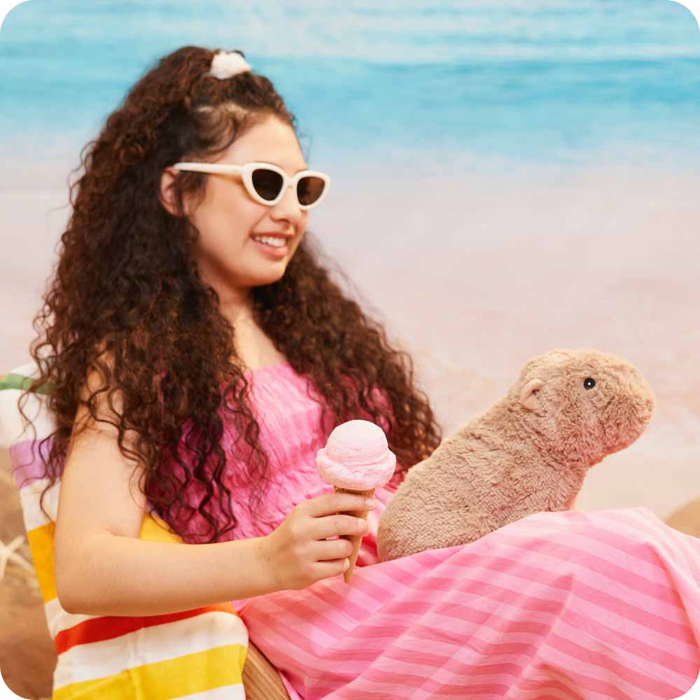 Woman in a pink dress with sunglasses holding an ice cream, sitting next to a plush groundhog on a beach.