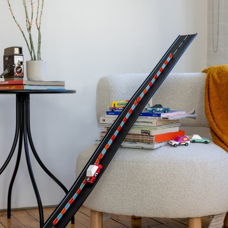 Model train track on a sofa with books and toy cars in a cozy living room setting.