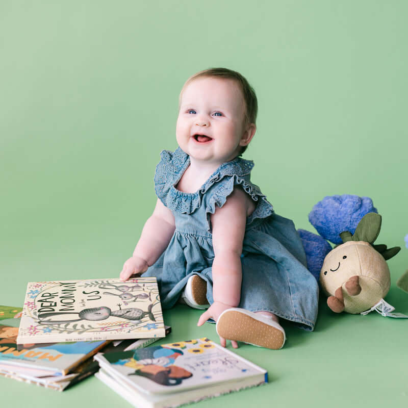 A baby girl sitting on a light green studio floor, wearing a denim blue ruffled dress. She is surrounded by children's books and a plush toy, looking up and smiling.