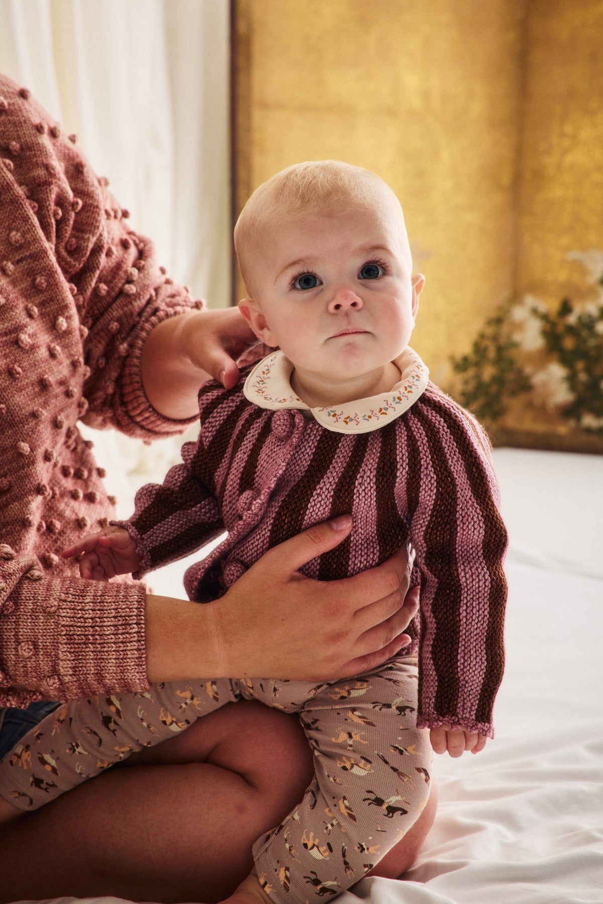 A baby wearing a knitted burgundy and pink striped sweater with a cream floral collar, sitting on a white bed while being held by an adult wearing a textured dusty rose sweater.