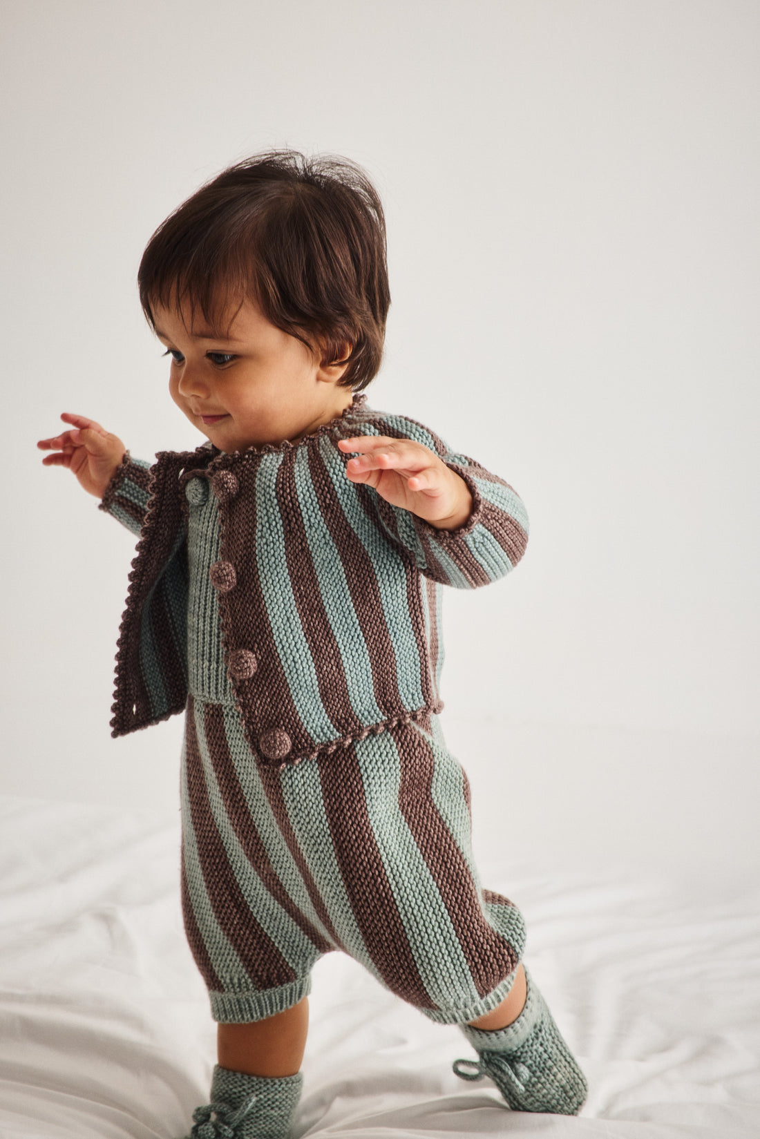 A toddler wearing a knitted striped romper in brown and sage green, standing on a white fabric surface against a light gray background.