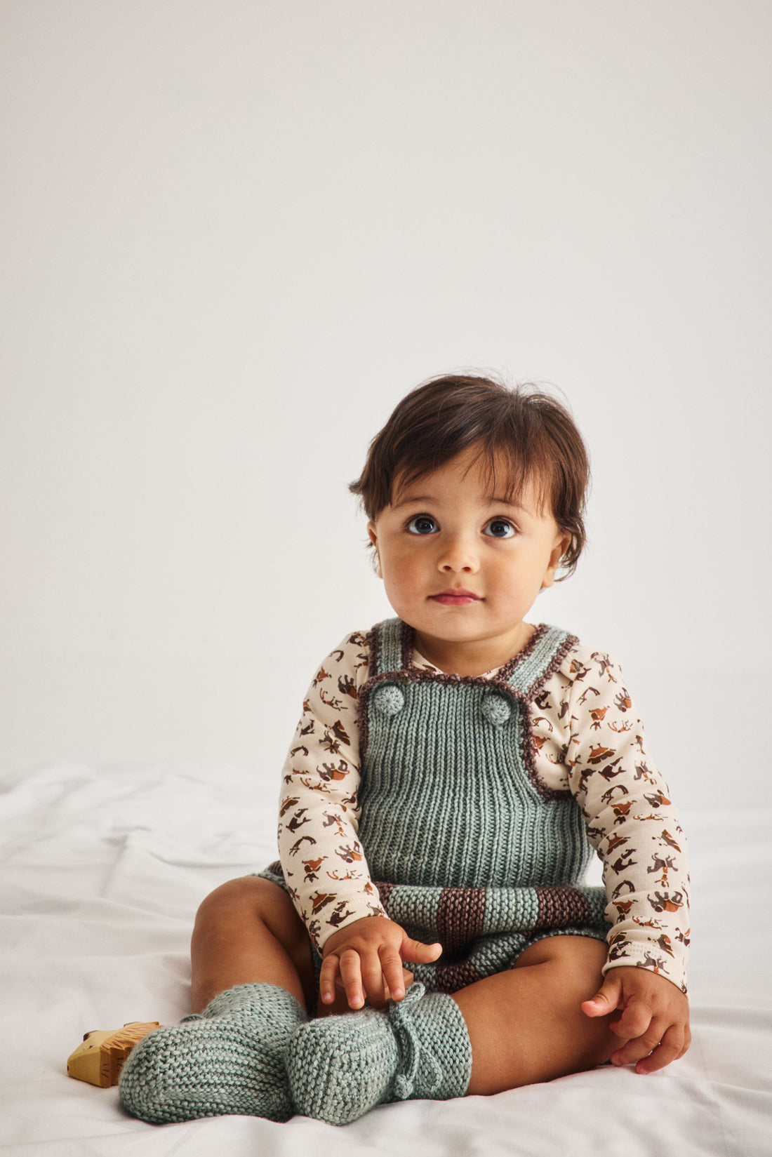 A toddler sitting on a white fabric surface wearing a knit overall romper in sage green with a brown and white patterned long-sleeve shirt underneath and matching knit booties.
