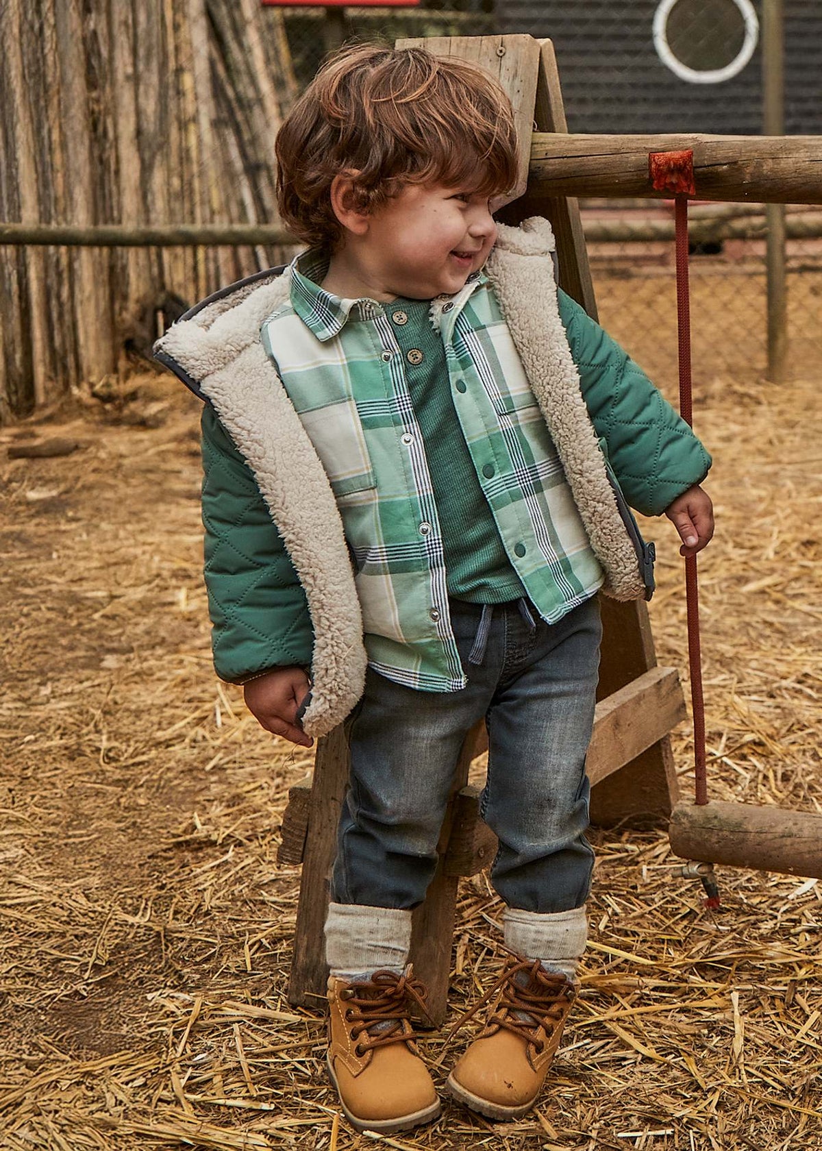 Child wearing a green plaid shirt, brown boots, and a puffer vest standing on straw.