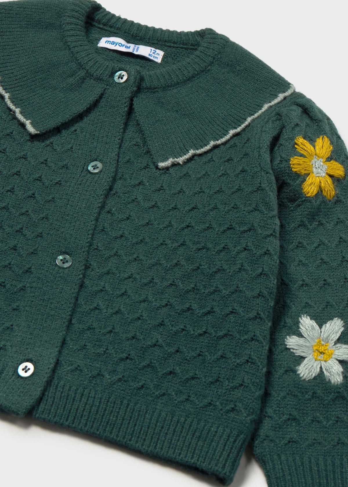 A close-up of a dark green knit cardigan with a textured waffle pattern, featuring embroidered yellow and white daisies on the sleeve and a matching green button closure.