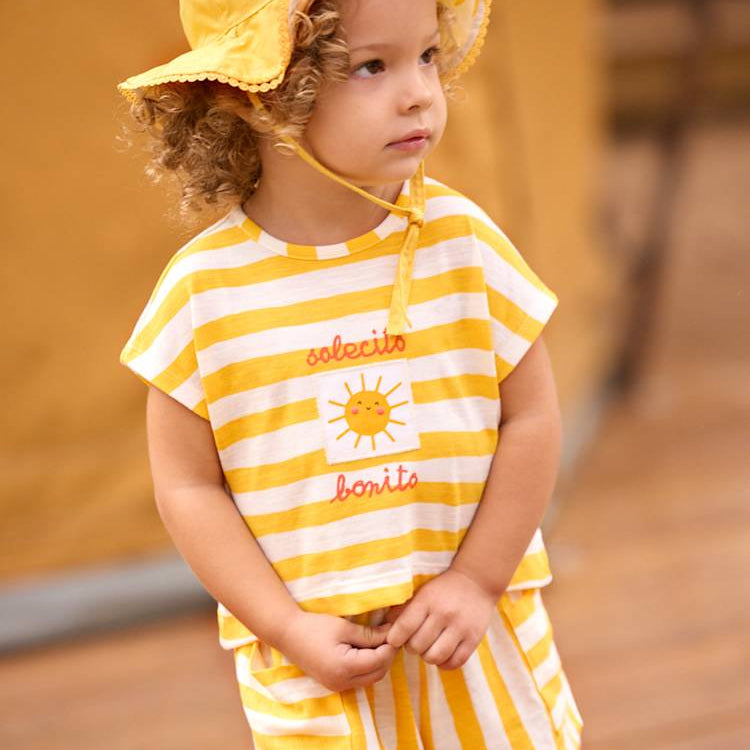 Child wearing a yellow and white striped outfit with a sun hat on a wooden floor.