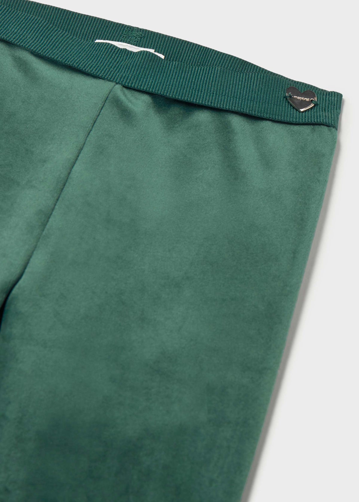 Close-up of green trousers showing the waistband with a white embroidered logo and a small metallic heart-shaped tag, on a light gray background.