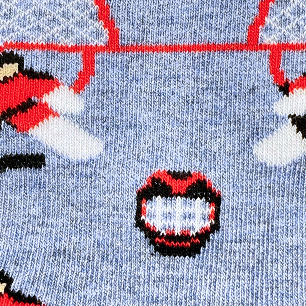 Close-up of a knit sock pattern on a heathered light blue background, featuring red, white, black, and beige graphic motifs including a stylized face and partial figures.