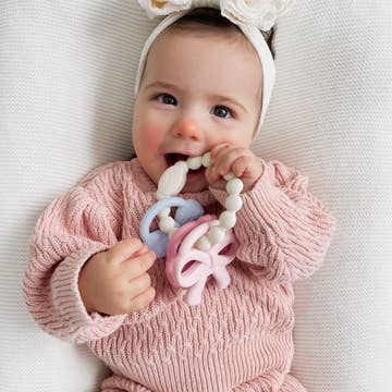 Baby in a pink outfit with a teething toy, lying on a white surface.
