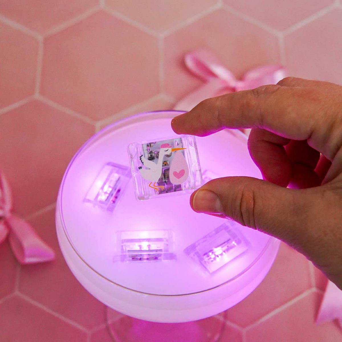 Hand interacting with a small round device emitting a pink glow on a pink tiled background.