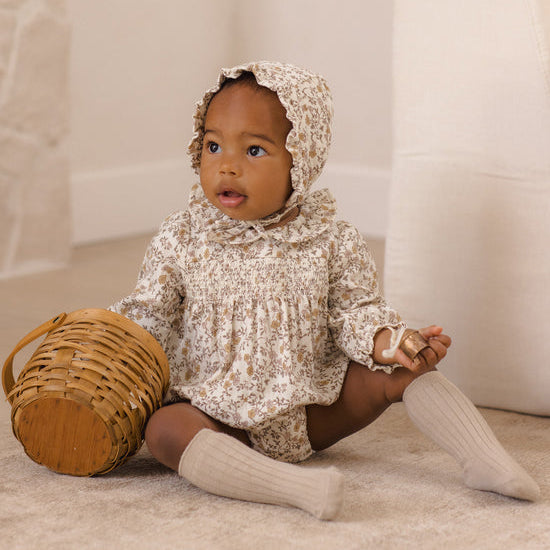 A baby sitting on a beige carpet wearing a cream and brown floral print smocked bodysuit with a matching bonnet, ribbed cream knee socks, and holding a small woven basket.