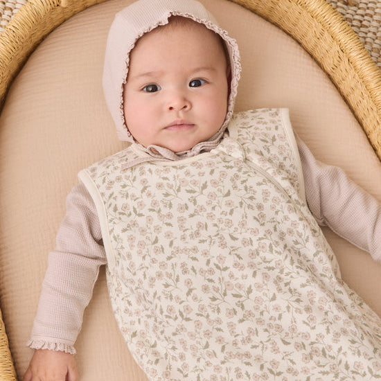 A baby wearing a cream floral-patterned sleeveless sleep sack over a light pink long-sleeve top and matching bonnet, lying on a beige cushion inside a woven rattan bassinet.