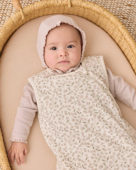 A baby wearing a cream floral-patterned sleeveless sleep sack over a light pink long-sleeve top and matching bonnet, lying on a beige cushion inside a woven rattan bassinet.