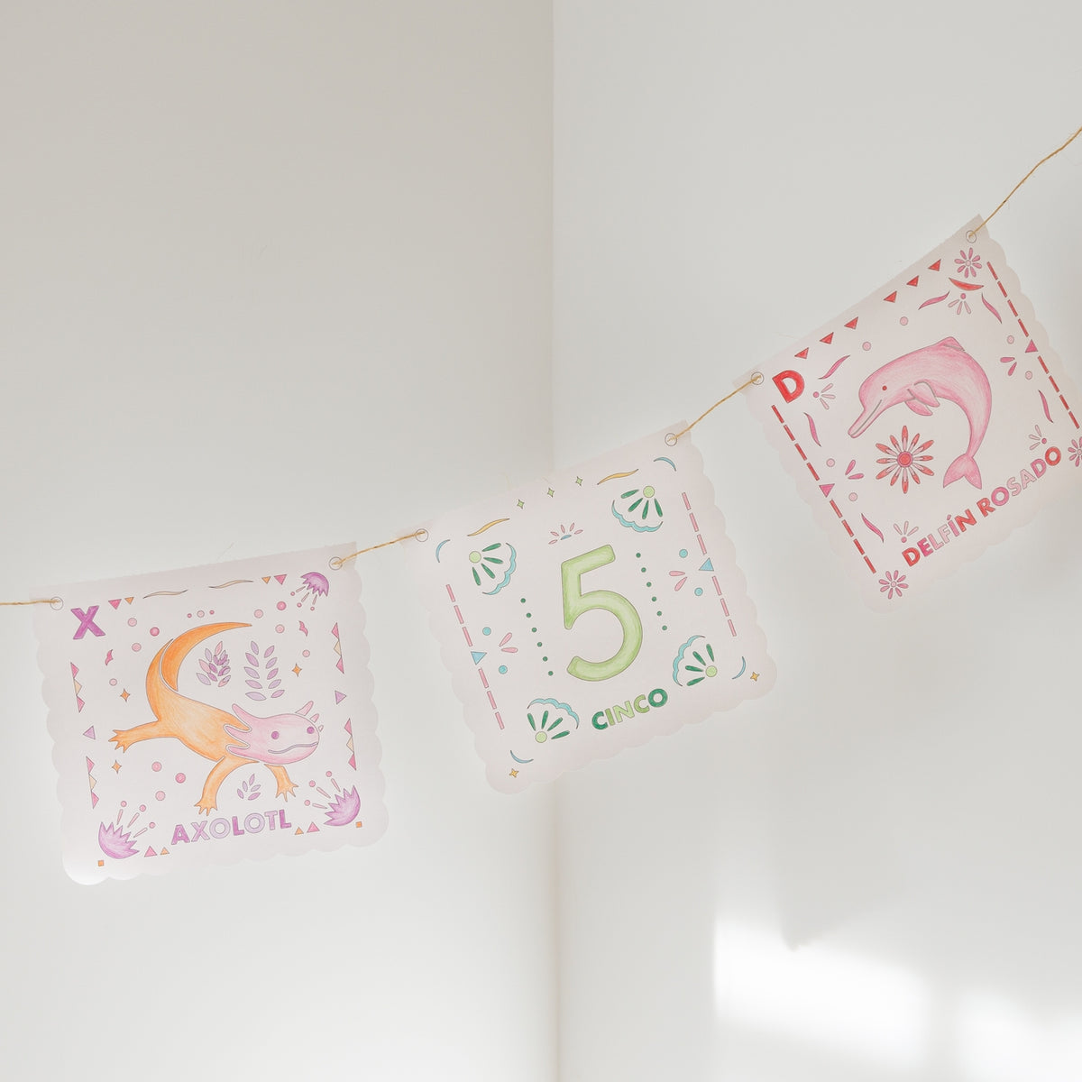 Three decorative square pennants strung along a thin cord, hanging against a white wall. The center pennant reads “5 Cinco” with green and orange lettering, while the side pennants feature pink and orange illustrations with Spanish text.