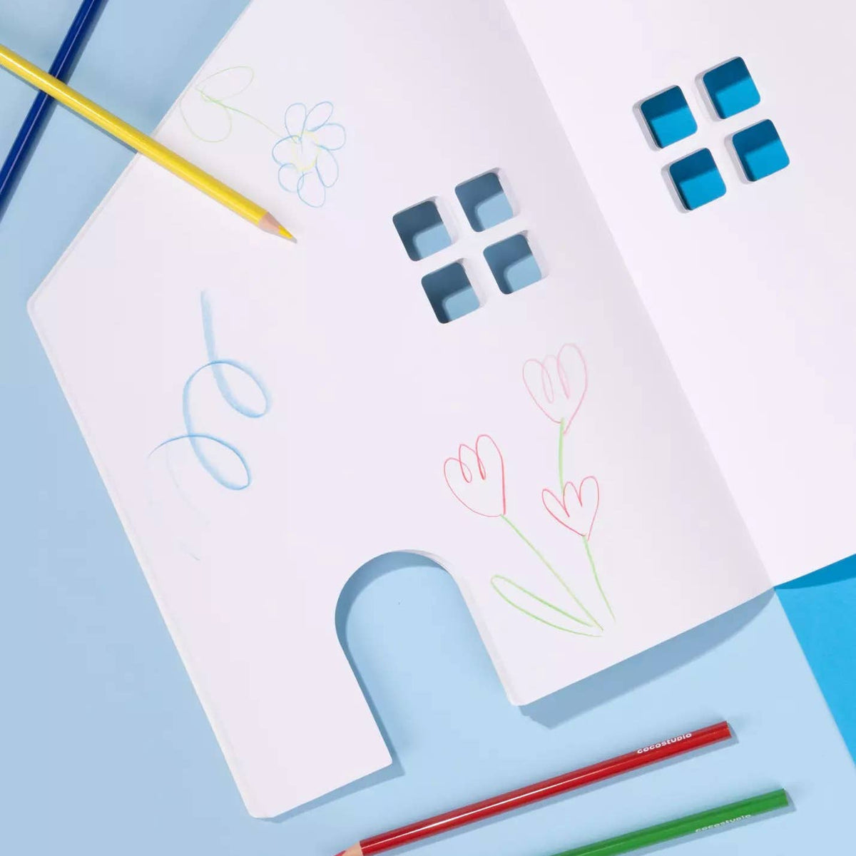 A white house-shaped children's drawing board with window and arch cutouts, shown on a light blue background with colored pencils around it. The board has simple hand-drawn flower and squiggle marks in blue, pink, green, and gray.
