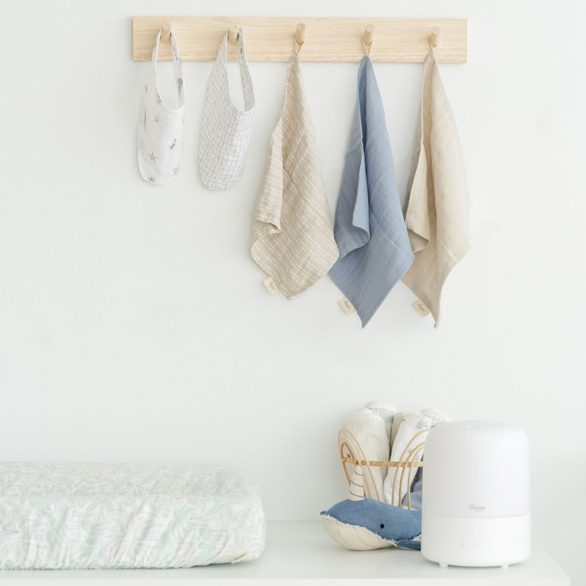 A light wood wall-mounted hook rack with four hanging fabric accessories in soft neutral and blue tones, displayed in a bright nursery-like setting above a changing surface with baby textiles and a white humidifier.