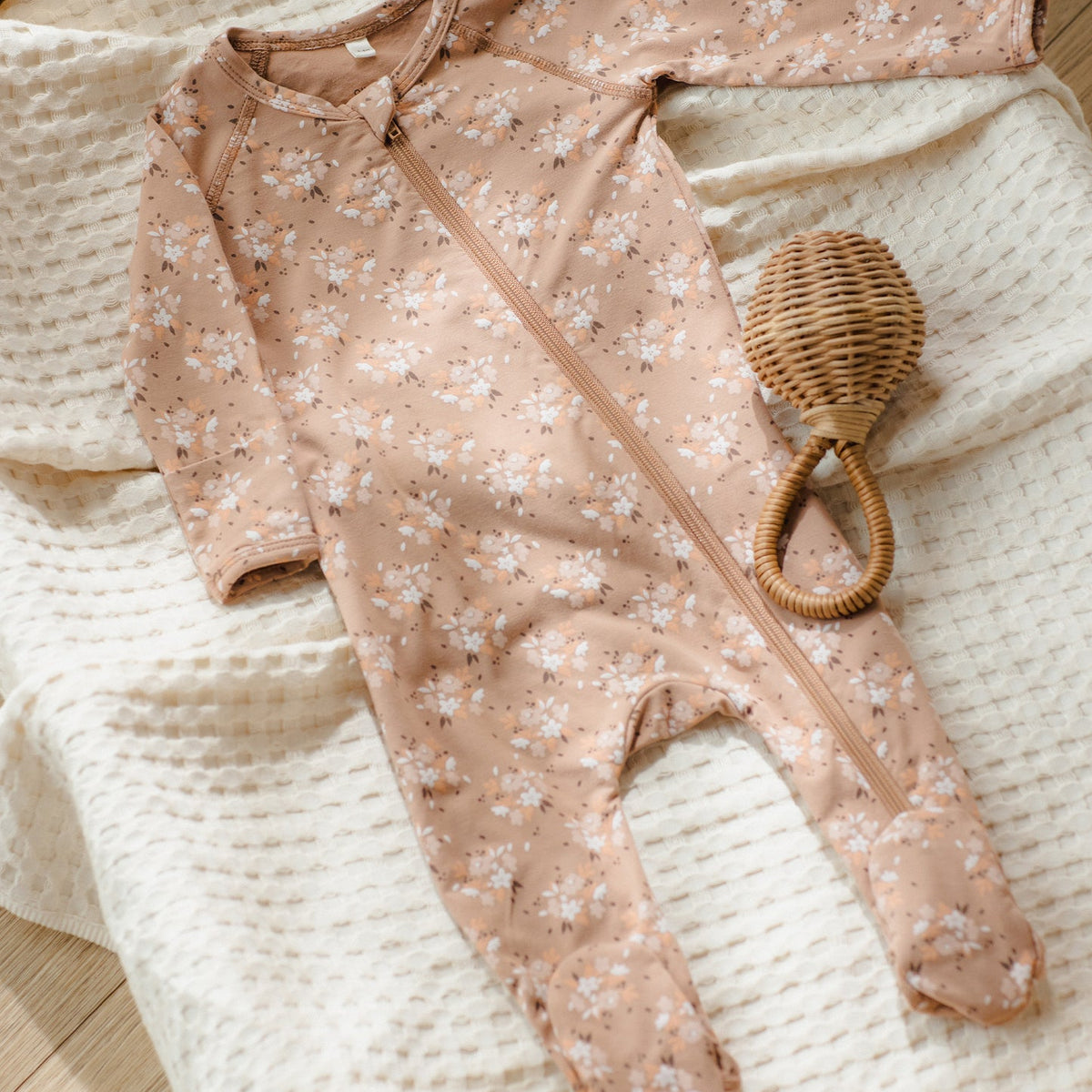 A baby footed sleeper in a dusty pink floral print with a front zipper, laid flat on a cream textured blanket next to a small woven rattleskin rattle.
