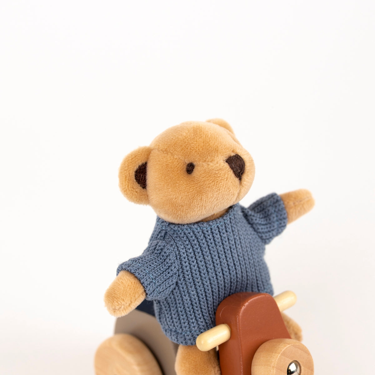 Teddy bear in a blue sweater sitting on a wooden toy with a plain background