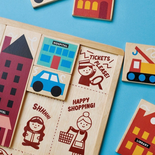 A close-up, top-down view of the Mentari Wooden Town Puzzle showing colorful, chunky wooden puzzle pieces partially placed in their tray, featuring buildings, a blue car, and illustrated characters with text reading "Tickets Please," "Shhhh!," and "Happy Shopping!"