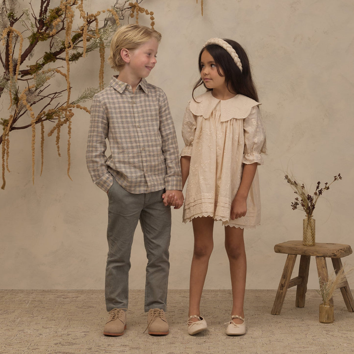 A young boy and girl standing hand-in-hand in a rustic studio setting. The boy wears a beige and brown plaid button-down shirt, grey trousers, and tan shoes.