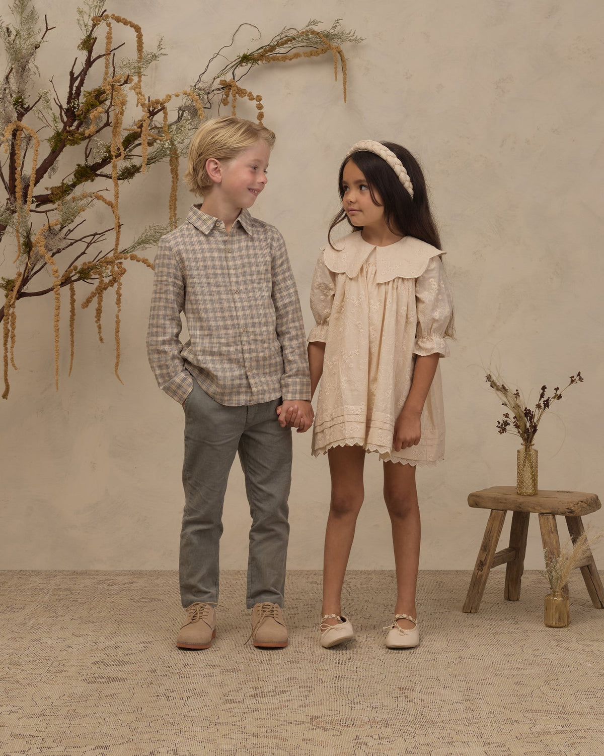 A young boy and girl standing hand-in-hand in a rustic studio setting. The boy wears a beige and brown plaid button-down shirt, grey trousers, and tan shoes.
