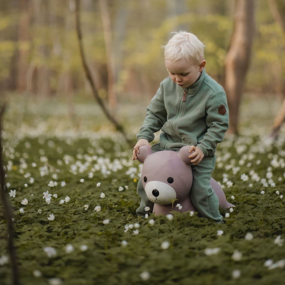 Child playing with a purple teddy bear in a forest with white flowers on the ground