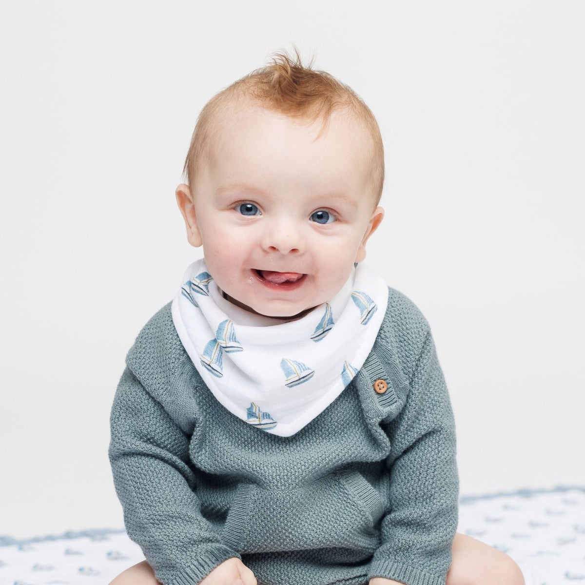 Baby wearing a green sweater and a white bib with blue sailboat pattern on a light gray background
