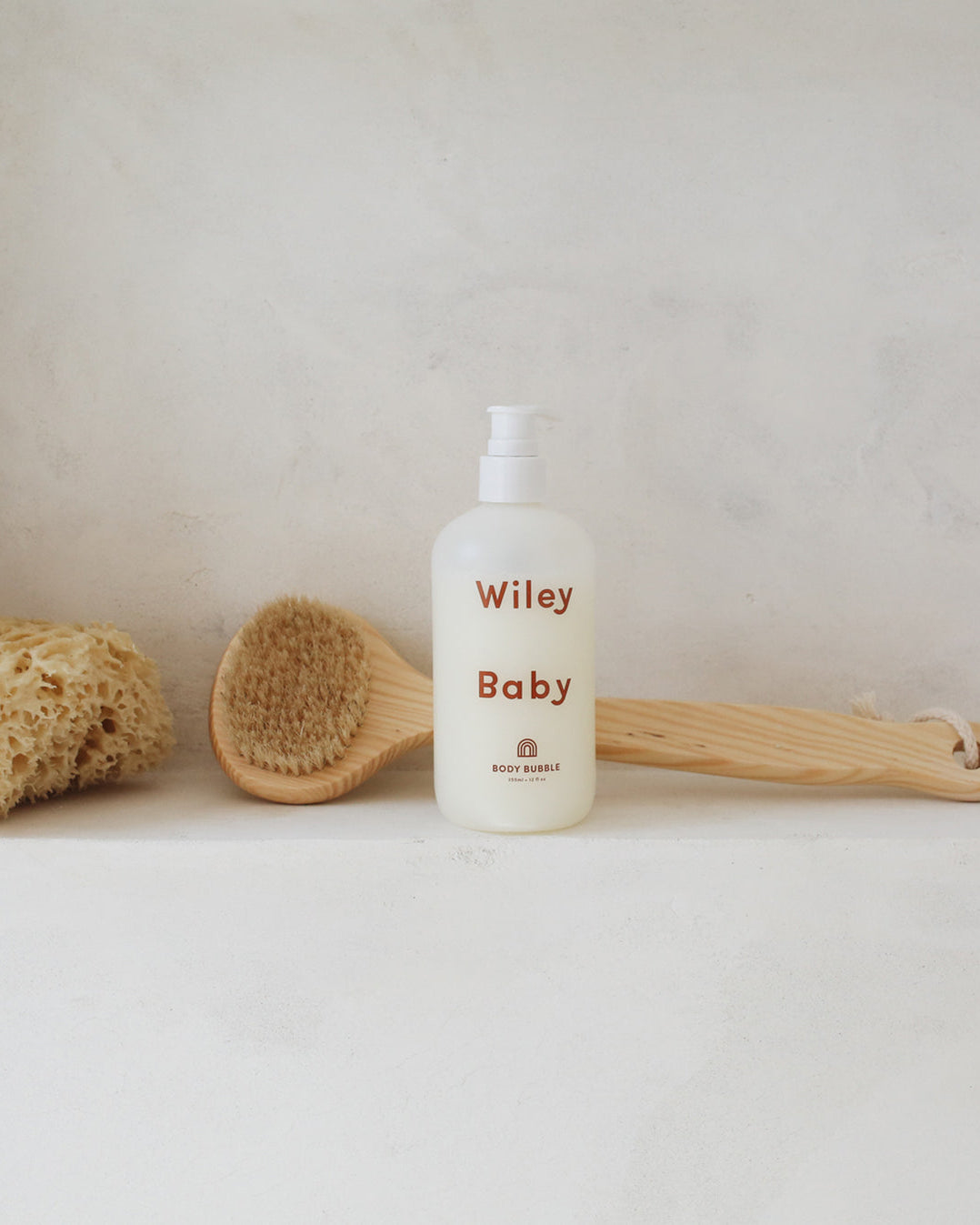 A bottle of Wiley Baby baby shampoo stands on a light surface against a textured off-white wall, styled with a wooden brush and a natural sponge.