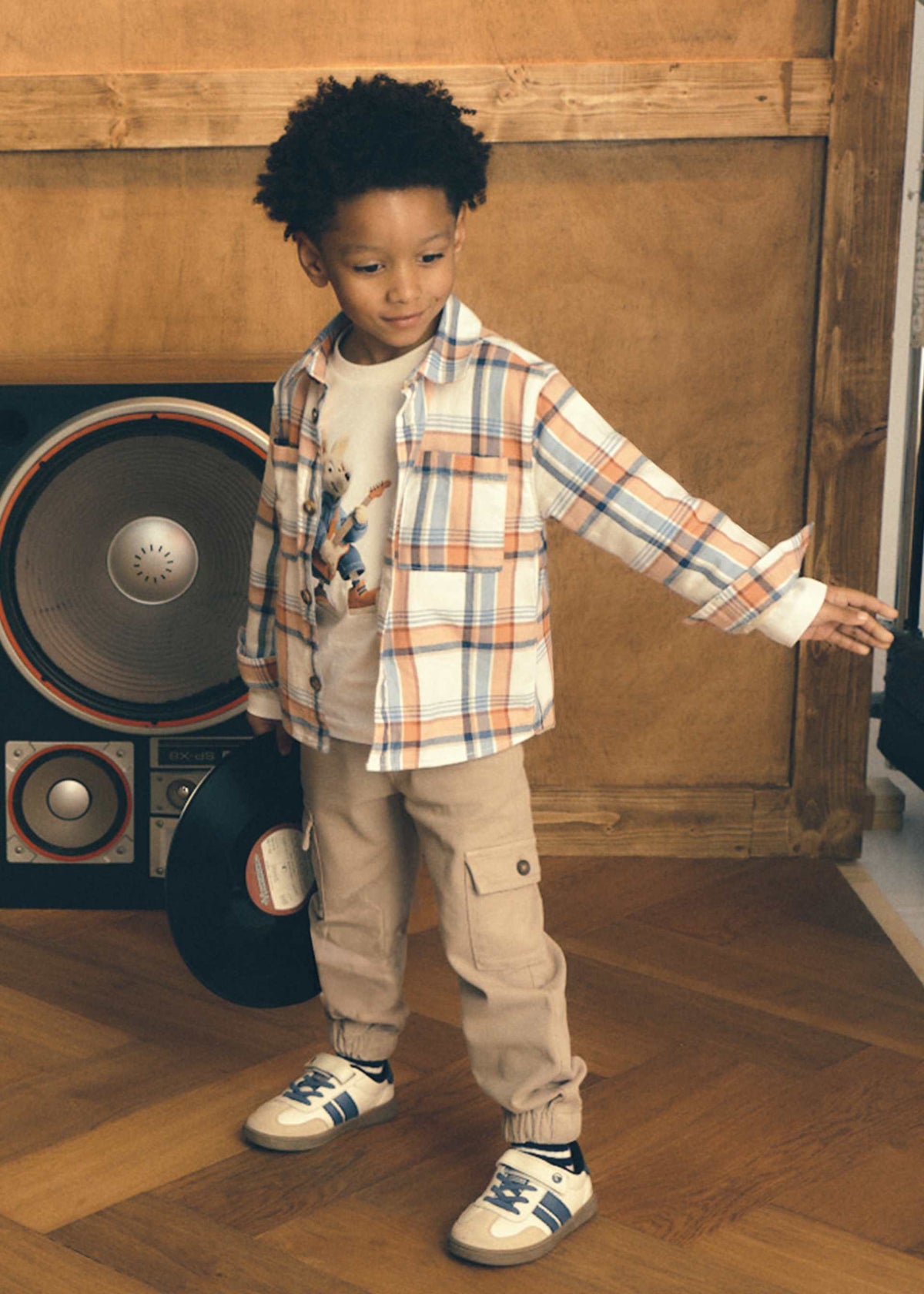 A young boy wearing a plaid button-up shirt, beige cargo pants, and white sneakers stands in a room with wood paneling and a large speaker behind him.