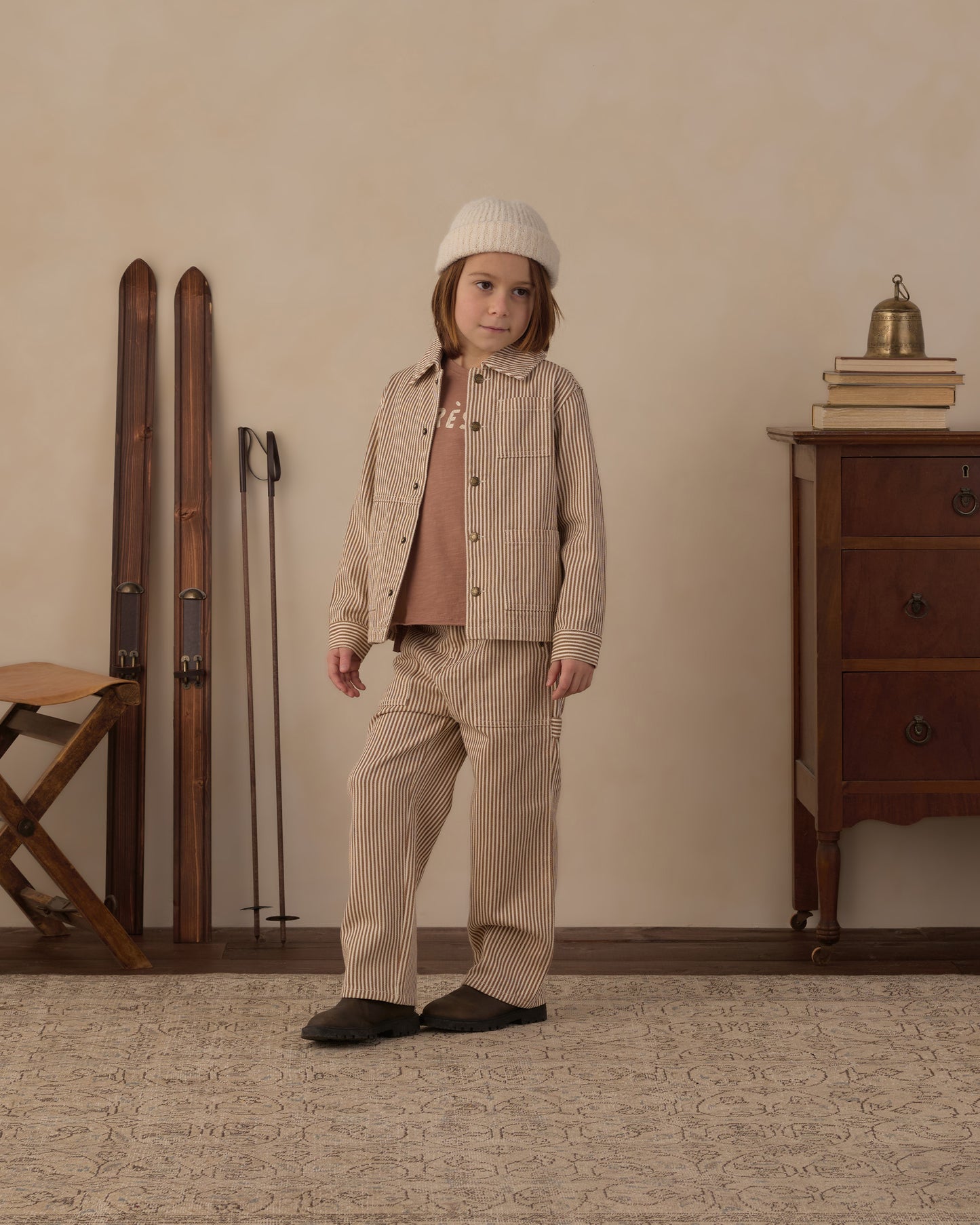 A child stands in a styled indoor room wearing a beige corduroy jacket and matching pants over a dusty rose top, with a cream knit beanie and dark slip-on shoes. Ski boots and poles lean against the wall on the left, and a wooden cabinet with books and a small brass bell sits on the right.