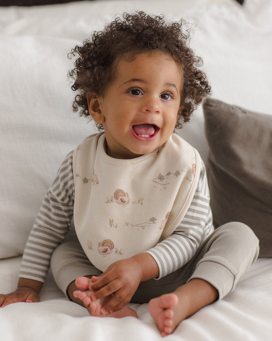 A smiling toddler with curly brown hair sits on a white bed wearing a cream bib with small illustrated motifs over a gray and white striped long-sleeve top and gray pants.