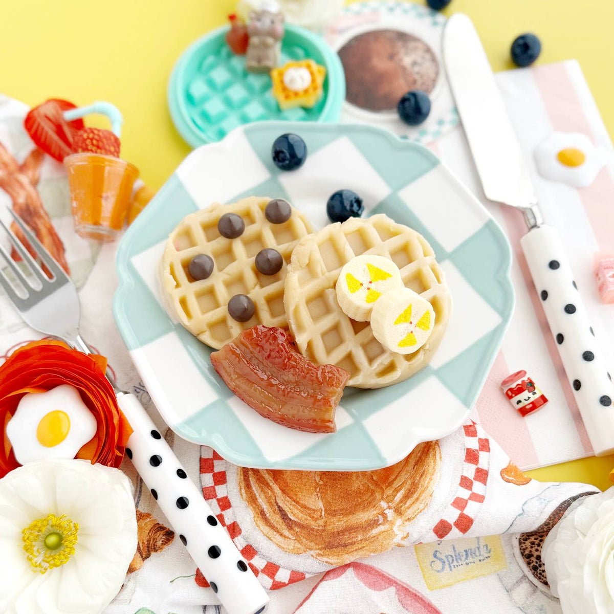 Miniature waffles with toppings on a checkered plate with utensils and decorative items.