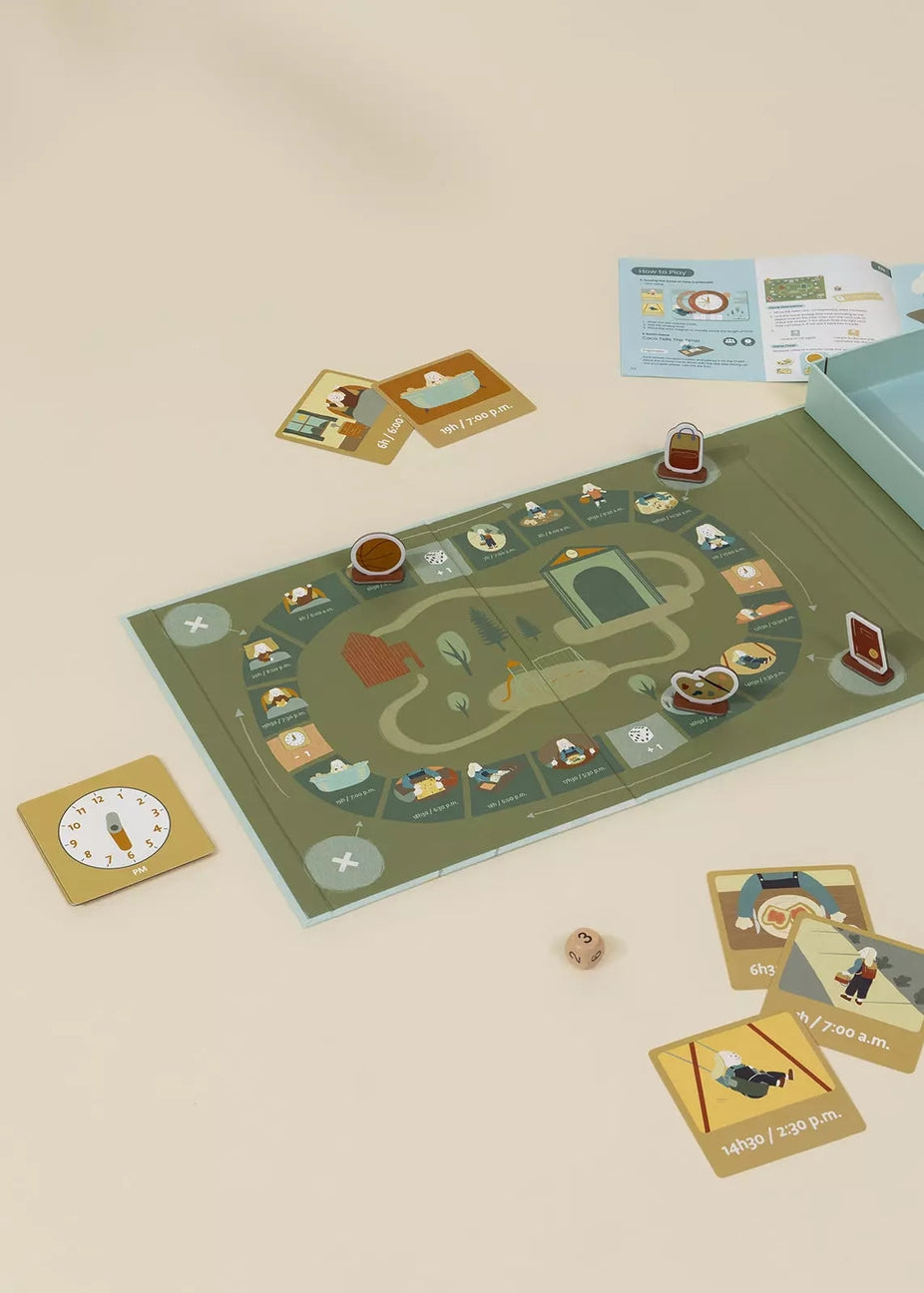 A board game set arranged on a warm beige surface, including a long illustrated game board, cards, a clock tile, dice, and a light blue storage box.
