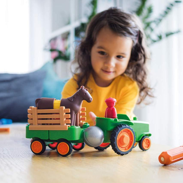 A young child smiling as they play on the floor with the green Smartmax My First Farm Tractor set, which features a brown toy horse pulling a cart and a red driver figure in the front.