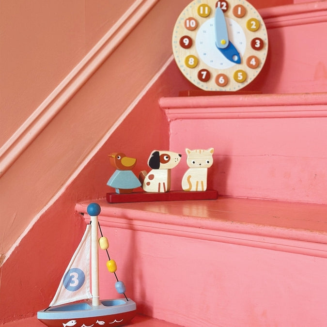 Wooden Pet Stacker toy by Mentari featuring a rectangular red base holding three removable pieces: a spotted white and brown puppy on the left, a blue and yellow bird on a tree in the center, and a white kitten with orange markings on the right, resting on a pink staircase beneath a wall-mounted blue and white clock.