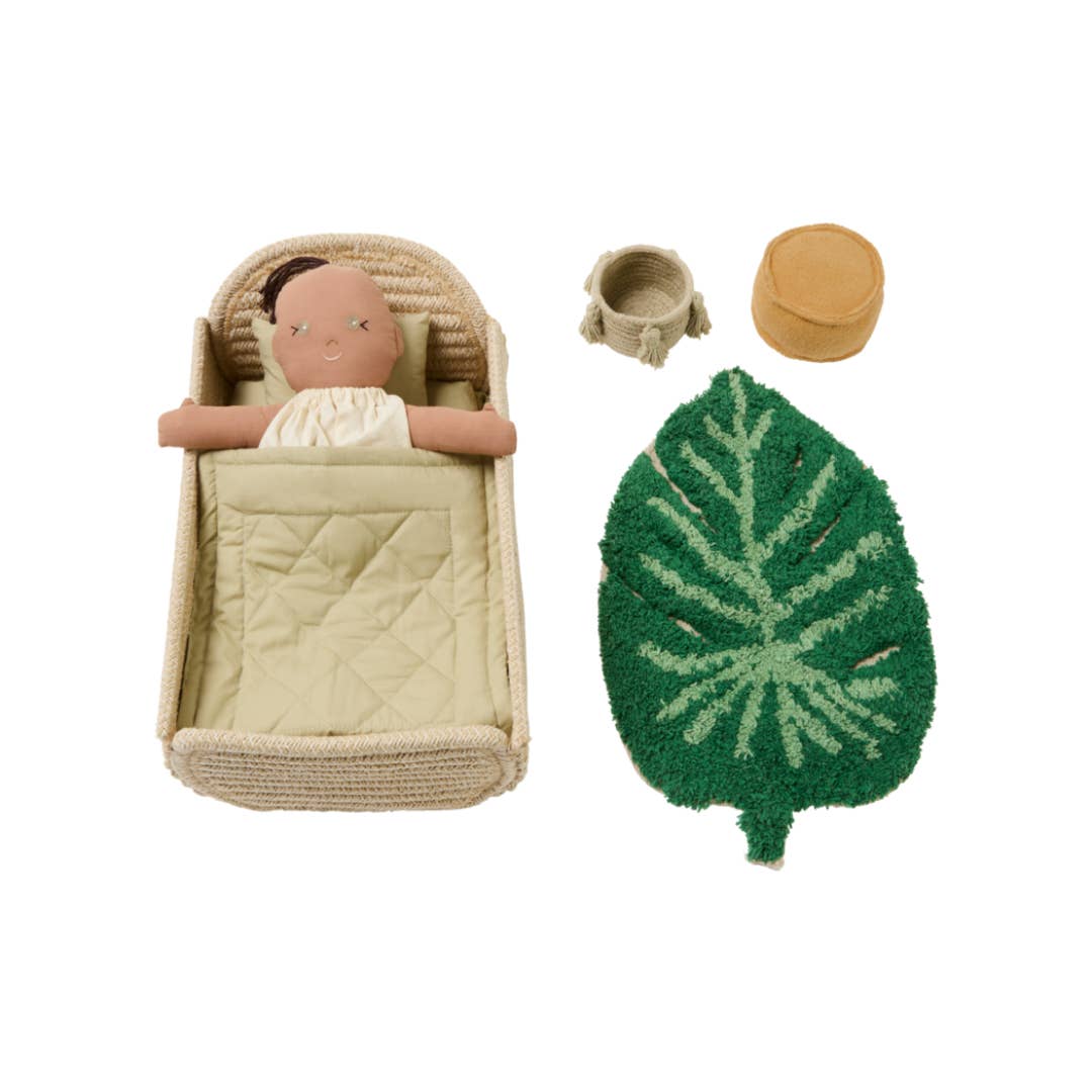 A doll play set featuring a woven bassinet with a quilted cream blanket, a doll with a pale face and white dress, a small woven basket, a round tan cup, and a green leaf-shaped rug with a pale green leaf pattern, all arranged on a white background.