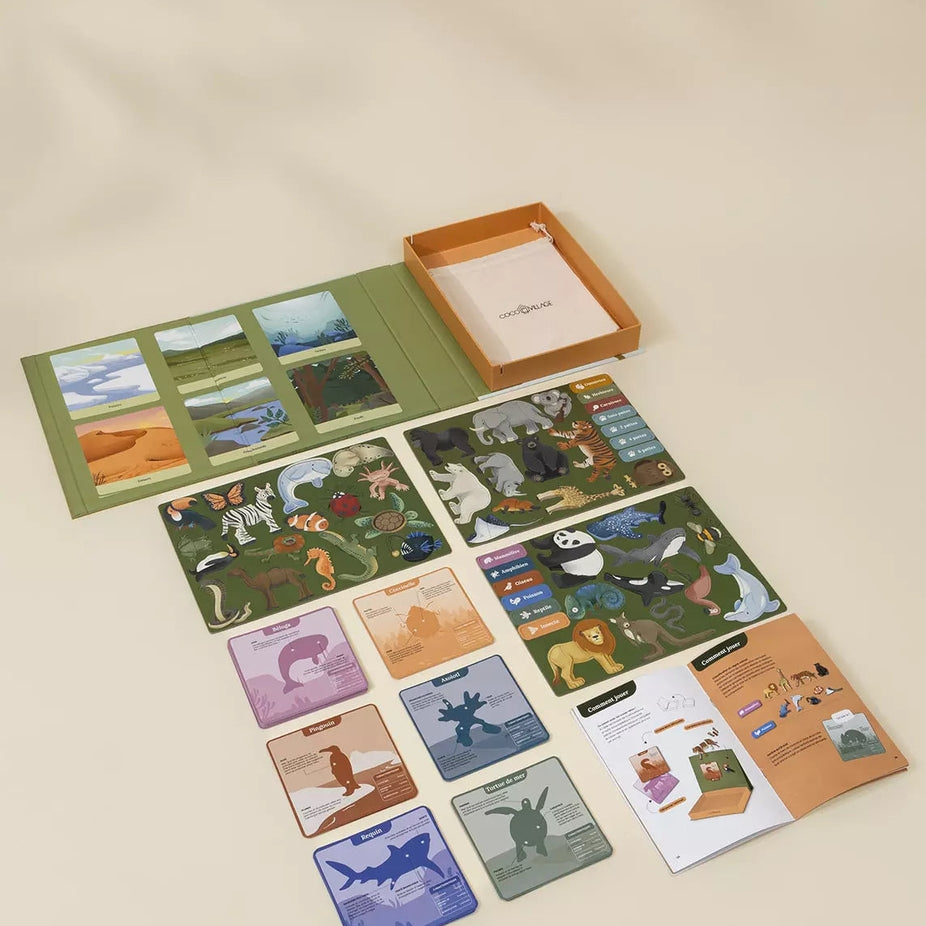 A children's card game set arranged on a warm beige background, including illustrated cards, a green play mat, an orange storage box, and instruction sheets.